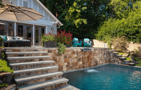 Wide stacked stone steps lead to the upper level of this custom backyard renovation featuring a modern pool house and natural paver patio with separate lounge areas