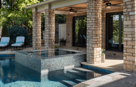 Large, stacked stone columns frame the flagstone covered porch and knife edge spa finished in captivating shades of light blue and dark blue 1” x 1” pool tiles. The infinity edge pool features a vibrant pool tile in Coral-Teal and a Black Marble PebbleTec finish.
