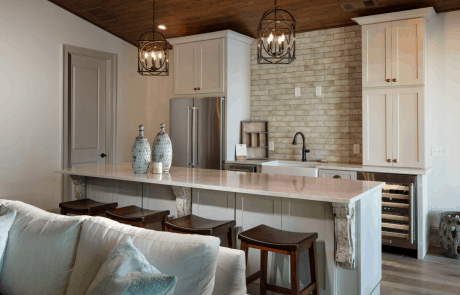 Detached custom pool house addition decorated in neutral tones features a vaulted cedar tongue & groove ceiling and full custom kitchen with bar island, white quartz countertops, soft white cabinetry, and a sand-colored matte porcelain subway tile backsplash. A set of modern dark bronze chandeliers and decorative antiqued corbels add a hint of luxury to the space.