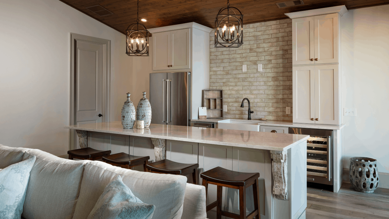 Detached custom pool house addition decorated in neutral tones features a vaulted cedar tongue & groove ceiling and full custom kitchen with bar island, white quartz countertops, soft white cabinetry, and a sand-colored matte porcelain subway tile backsplash. A set of modern dark bronze chandeliers and decorative antiqued corbels add a hint of luxury to the space.