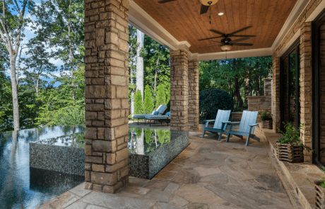 Classic style covered flagstone patio features a cedar tongue and groove ceiling, and unobstructed water views of the lake. Large, stacked stone columns frame the sleek knife edge spa finished in captivating shades of light blue and dark blue 1” x 1” pool tiles with the dramatic vanishing edge pool blending into the horizon.