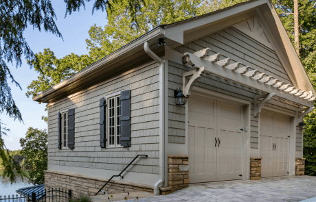 Custom two car garage addition sits on the level above the pool house and features a gabled roof, cedar shake exterior with stacked stone accents, an eyebrow pergola, black custom shutters, and aluminum gutters. The heated driveway is finished in Belgard’s “Avondale” Lafitt pavers.