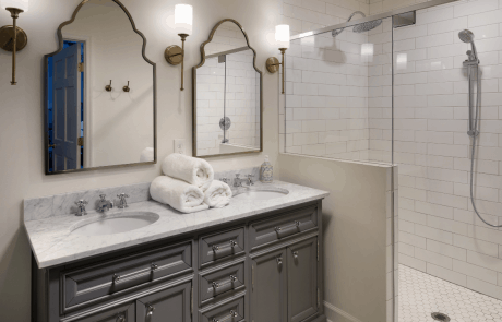 This transitional gray and white master bath remodel includes a large walk-in shower with glass hinged door and white subway tile walls, a double sink vanity paired with gray shaker cabinets and white marble countertops and a matte white hexagon floor tile. The brass sconces and vintage arched mirrors complete the look of this master bath remodel.