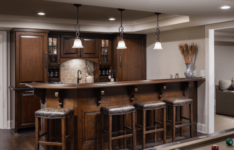 Open concept basement remodel includes a custom bar with cherry wood shaker cabinets and glass doors with antique glass, brick backsplash, stainless steel appliances, brown leather bar stools, and traditional accents and light fixtures.