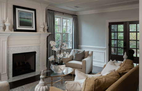 Basement renovation features an elegant sitting room with a white marble fireplace and custom mill-work trim, neutral color furnishings and an oval glass top coffee table with gold accents.