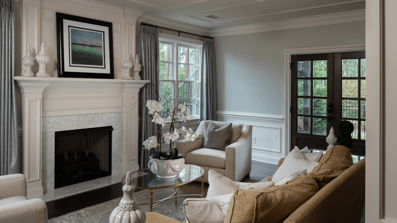 Basement renovation features an elegant sitting room with a white marble fireplace and custom mill-work trim, neutral color furnishings and an oval glass top coffee table with gold accents.