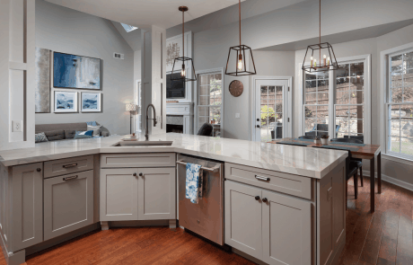 Contemporary shaker kitchen remodel features an open concept L-shaped kitchen island with natural quartz countertops, classic gray shaker cabinets, graphite colored hardware, dark bronze pendant lights and brown wood floors overlooking the eat in kitchen area and family room.
