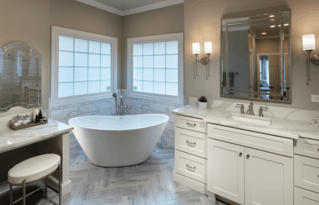 This modern master bath remodel includes separate built-in vanities with white shaker cabinets, brushed nickel hardware, white and gray quartz countertops and white rectangular under mount sinks. The silver luxe vanity mirror and 2-light sconces in antique nickel add a touch of sophistication to the space. Other features include a separate make-up vanity with arched mirror, white freestanding tub with brushed nickel tub filler & hand shower, a silver porcelain tile floor and coffered ceiling.