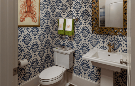 This transitional blue and white powder room is clad in geometric wallpaper and boasts a gold-finish hanging mirror, and a rectangular white pedestal sink with a brass faucet. Dark hardwood floors, and a bold orange Octopus print, and accent towels provide pops of color to the finished bathroom.