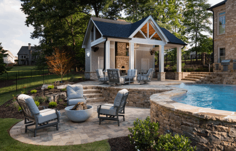 Custom pool design and patio design in Atlanta includes Belgard’s "Lafitt Avondale" paver patios with a stacked stone raised beam and stone steps. The lower patio features a separate firepit area and modern wood burning fire bowl. A traditional style pool house and open-air pool cabana features a gabled roof, Hardie-Shake siding, a custom outdoor stacked stone fireplace with pizza oven, covered dining area, private bathroom, and storage room.