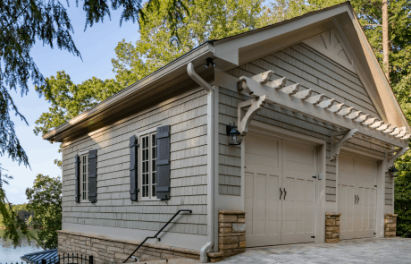 Custom two car garage addition sits on the level above the pool house and features a gabled roof, cedar shake exterior with stacked stone accents, black custom shutters, and aluminum gutters. The heated driveway is finished in Belgard’s “Avondale” Lafitt pavers.