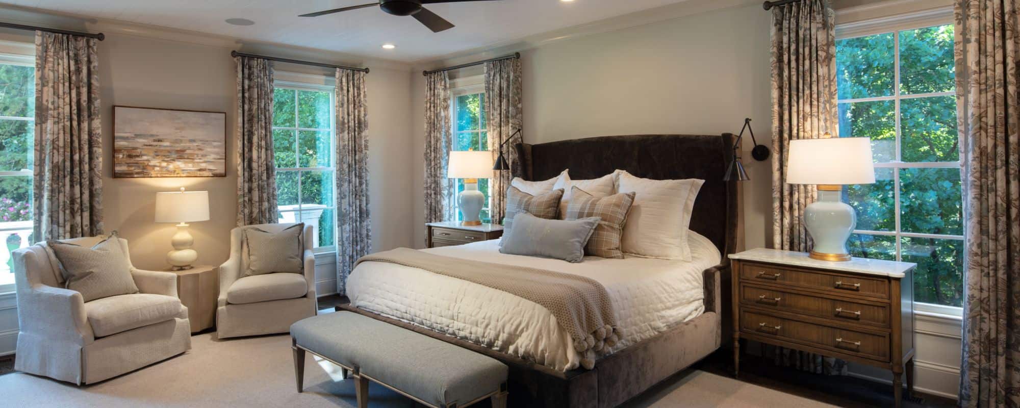 Neutral colored large master bedroom is painted in Benjamin Moore’s Edgecomb Gray with custom trim work, white painted tongue-and-groove ceiling, secret hidden door, dark bronze wall sconces and ceiling fan, brown hardwood floors and designer finishes.