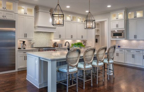 Large, transitional style kitchen with white honed quartzite countertops, oversized kitchen island, white shaker cabinets with black hardware, Sub-Zero appliances, penny mosaic backsplash, gold and black lantern pendants, and dark brown hardwood floors.