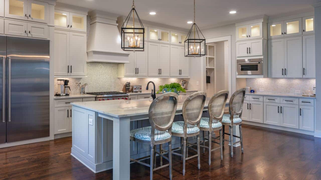 Large, transitional style kitchen with white honed quartzite countertops, oversized kitchen island, white shaker cabinets with black hardware, Sub-Zero appliances, penny mosaic backsplash, gold and black lantern pendants, and dark brown hardwood floors.