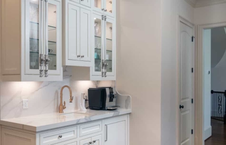Separate custom beverage & coffee station features undermount sink with Waterstone pull down faucet in Satin Brass, white custom inset cabinets with decorative glass doors and brass Cremone bolt hardware, Kalos Bianco porcelain countertops, porcelain slab backsplash and undercabinet lighting.