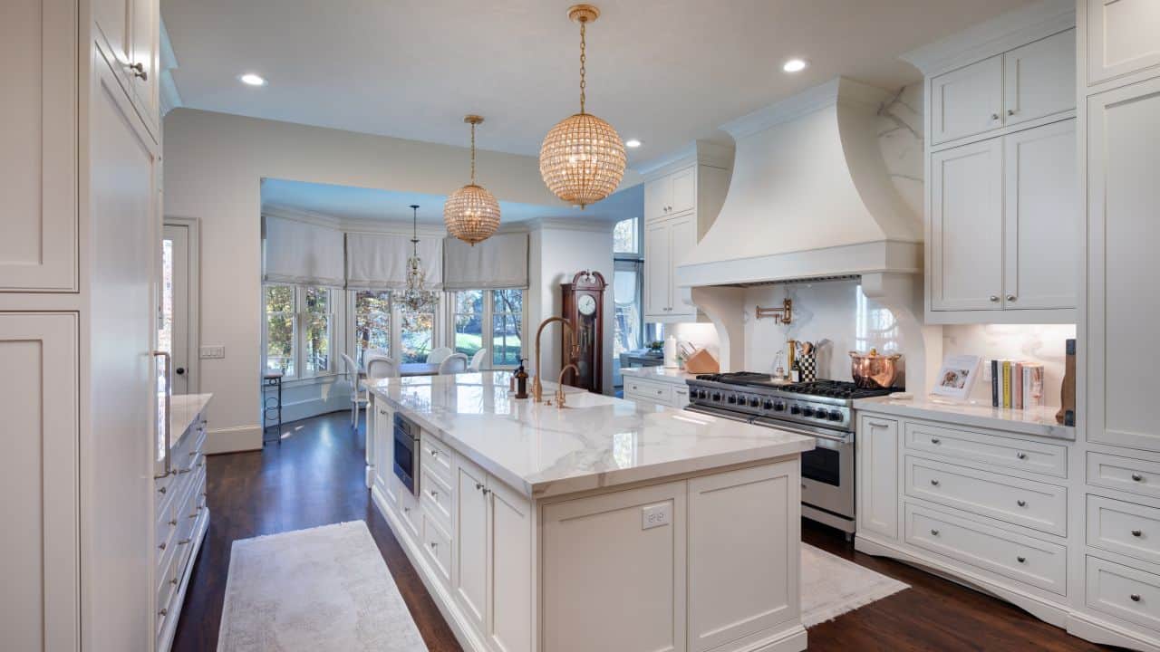 Custom kitchen with Polished Kalos Bianco Porcelain Countertops, Kohler Apron Sink, Waterstone Luxury Faucet in Antique Brass, White Custom Inset Cabinetry, Antique Brass Crystal Chandelier Pendants, 60” Viking Dual Range Custom Vent Hood. Oversized Island. Porcelain Slab Backsplash, and Dark Brown Hardwood Floors