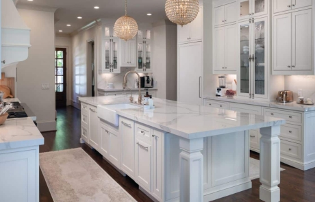 Custom Kitchen Remodel With White Custom Inset Cabinets with Decorative Glass Doors, Polished Kalos Bianco Porcelain Countertops, Kohler Apron Sink, Waterstone Luxury Faucet in Antique Brass, Antique Brass Crystal Chandelier Pendants, Oversized Island, Undercabinet lighting, and Brass Cremone Bolt Hardware