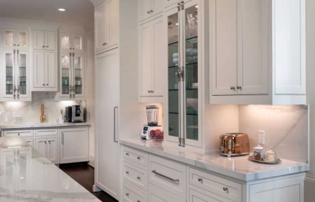 Custom kitchen remodel features white custom inset cabinets with decorative glass doors, custom appliance panels, polished Kalos Bianco porcelain countertops, porcelain slab backsplash, oversized island, undercabinet lighting, separate beverage/coffee station with undermount bar sink and Waterstone pull down faucet in Satin Brass.
