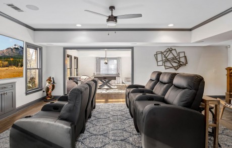 This transitional open concept basement renovation includes a family room and home theater furnished with luxurious gray leather stadium seating, a large flat screen TV and a behind the couch bar table with stool seating.