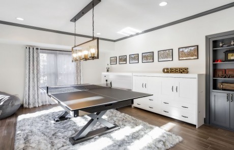 Large basement game room remodel features a set of hidden horizontal murphy bed wall units in white, dark gray built-in book shelves, and a ping pong table with black modern linear pendant light above.