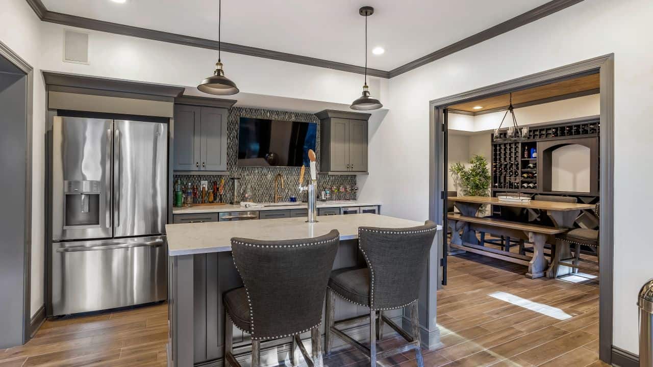This large basement remodel features a fully equipped contemporary shaker kitchen and bar with grey shaker cabinets, gray and white glass mosaic backsplash, white Viatera Minuet quartz countertops, stainless steel appliances including a built-in kegerator and light brown porcelain plank tile flooring.