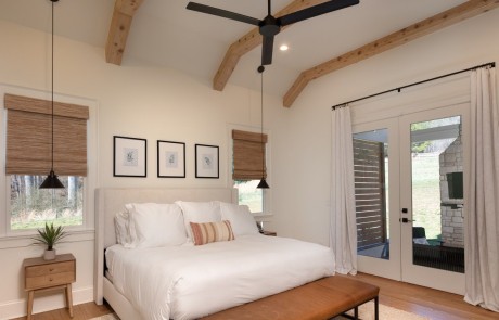 This white master bedroom includes a cathedral ceiling with decorative cedar beams in a natural wood tone, modern black hanging pendant lights, and large double doors with private access to an outdoor covered sitting area and outdoor fireplace.