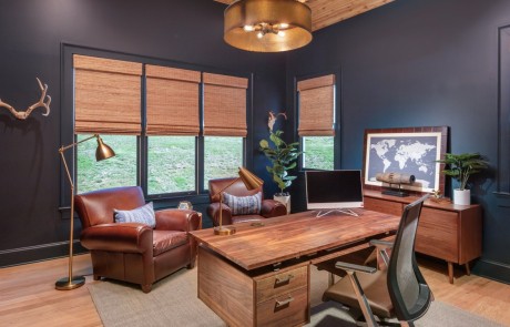The modern rustic design of this home office has a masculine moody vibe with a rough-sawn cedar ceiling, light oak hardwood floors, dark blue walls, and brown leather arm chairs.