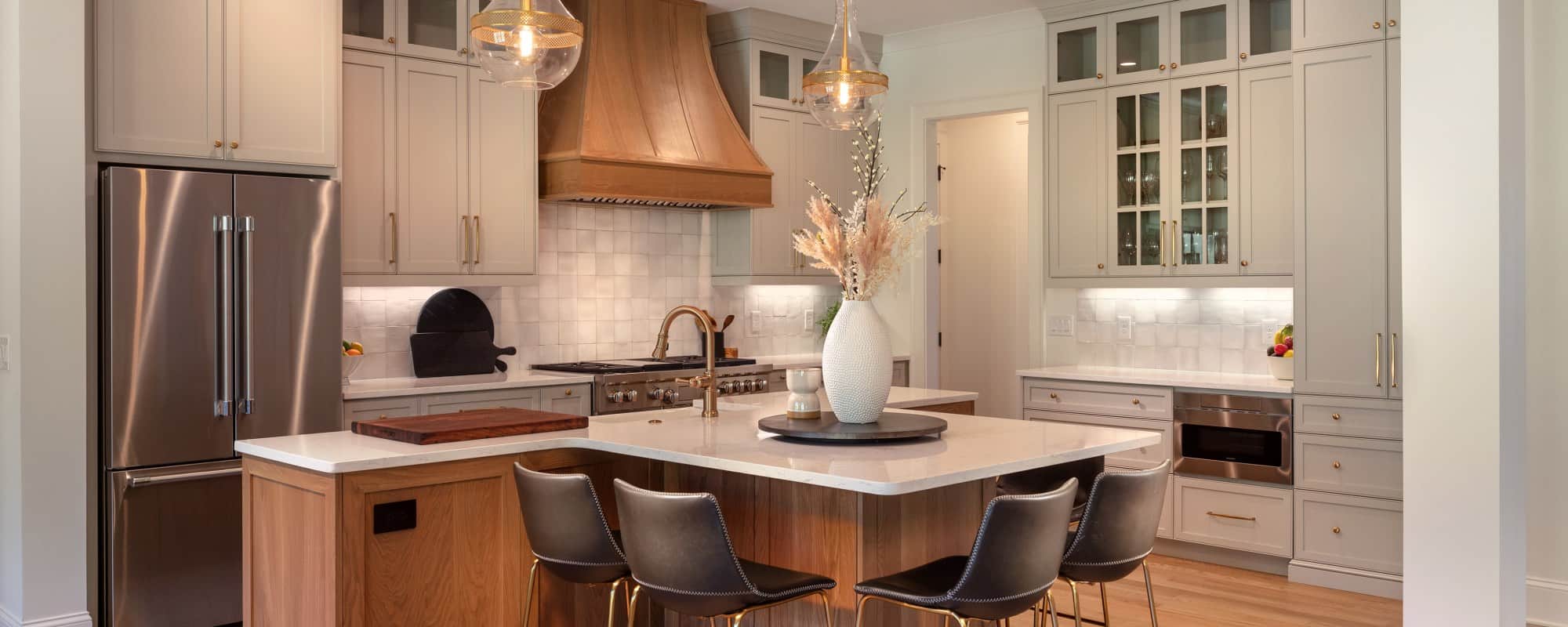 Large open-concept kitchen features a 48” Wolf Dual Fuel range with a custom wood vent hood, white Quartz countertops, farmhouse sink, white apron sink, Zellige square tile backsplash, gray shaker cabinets with gold hardware and light oak hardwood floors. An oversized T-shaped island provides seating for six.