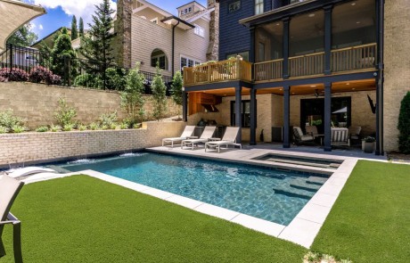 Contemporary backyard design features a custom cocktail pool and flush spa with a Baja shelf, pool loungers, and an eye-catching raised wall of sheer descent waterfalls. The modern, clean lines of the light-colored concrete patio and pool deck contrast beautifully with the artificial turf in this luxurious custom outdoor living space.