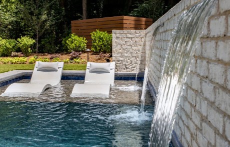 Custom tanning ledge with stylish white pool chaise loungers and a raised brick wall of sheer descent waterfalls provide the ultimate setting for relaxation in this contemporary pool design.