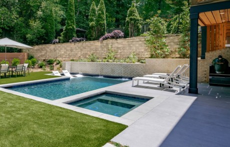 Custom designed modern plunge pool and flush spa features a large tanning ledge with loungers, a seating bench that runs the entire width of the pool and a feature wall of sheer descent waterfalls. The green artificial turf pops against the decorative concrete coping and pool deck, adding a clean, minimalistic feel to this contemporary backyard renovation.