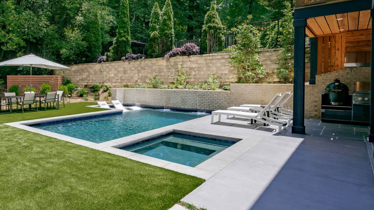 Custom designed modern plunge pool and flush spa features a large tanning ledge with loungers, a seating bench that runs the entire width of the pool and a feature wall of sheer descent waterfalls. The green artificial turf pops against the decorative concrete coping and pool deck, adding a clean, minimalistic feel to this contemporary backyard renovation.