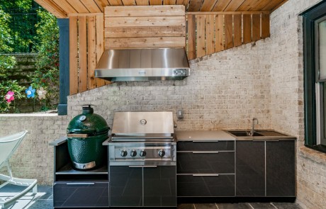 This modern outdoor kitchen design features dark gray aluminum outdoor cabinets, smooth concrete countertops, a stainless-steel vent hood with warm wood accents, built-in grill, sink, storage with cabinets and drawers and a Big Green Egg.