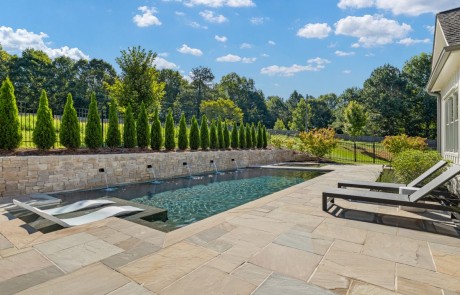 High end pool construction includes a sleek rectangular pool with a knife-edge perimeter overflow spa and tanning ledge. Surrounding the pool’s perimeter is a limestone stacked stone wall with decorative matte black wall scuppers and a light-colored Sandstone paver patio.