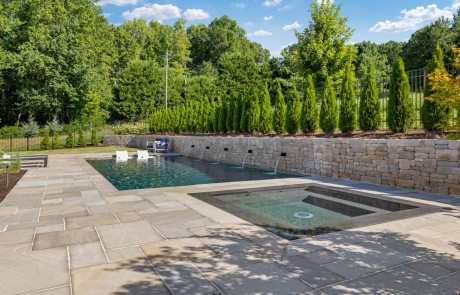 Exclusive pool design with contemporary style pool and knife-edge perimeter overflow spa finished in a Black Onyx Pebble Tec contrasts beautifully with the Sunset Sandstone paver patio, Indiana limestone stacked stone wall and lush landscaping.