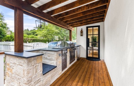 Custom-built outdoor kitchen finished in Texas Lueders limestone with stainless steel appliances and sleek black pearl leathered granite countertops sits beneath a modern cedar pergola finished in a dark oak stain.