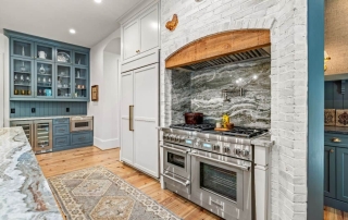 Luxury kitchen remodel features two-toned cabinets with custom glass-fronts and built-in storage solutions, lime-washed brick accent wall, wood beam range hood, statement kitchen backsplash, 48-Inch Dual-Fuel Pro Grand® Range, and panel ready appliances.